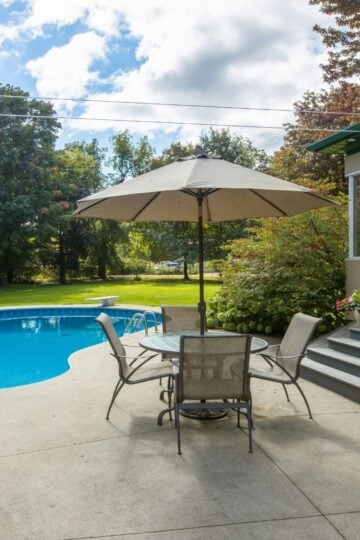 A garden with a round swimming pool featuring warmer pools for comfort, a concrete patio, table with four chairs and a large parasol, next to a house with steps, surrounded by trees and greenery.