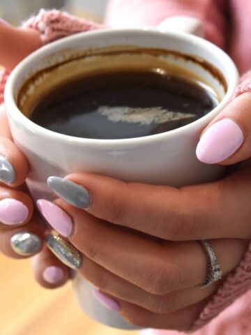 Person with pink and grey manicured nails holding a white mug filled with black coffee, drinking caffeine while wearing a pink jumper and a ring on one finger.
