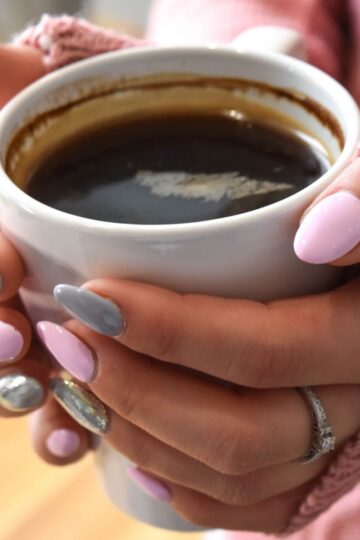 Person with pink and grey manicured nails holding a white mug filled with black coffee, drinking caffeine while wearing a pink jumper and a ring on one finger.