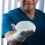 A person wearing blue scrubs and a white glove holds a silicone breast implant in their outstretched hand, highlighting considerations for breast implant replacement and the typical breast implant lifespan.