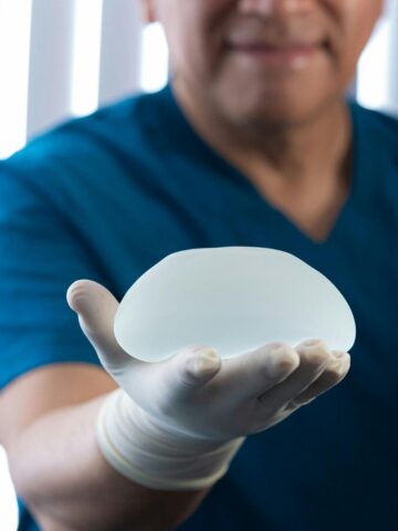 How Long Do Breast Implants Last and When Should They Be Replaced A person wearing blue scrubs and a white glove holds a silicone breast implant in their outstretched hand, highlighting considerations for breast implant replacement and the typical breast implant lifespan.