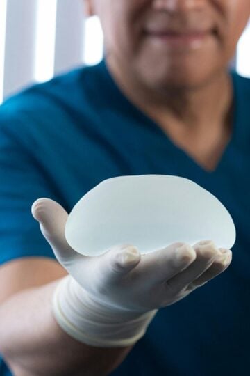 A person wearing blue scrubs and a white glove holds a silicone breast implant in their outstretched hand, highlighting considerations for breast implant replacement and the typical breast implant lifespan.