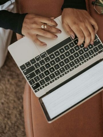 Person typing on a laptop placed on a brown surface, with glasses resting nearby. Keyboard and part of the screen displaying an Information Included section for a legitimate ESA Letter are visible.