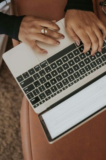 Person typing on a laptop placed on a brown surface, with glasses resting nearby. Keyboard and part of the screen displaying an Information Included section for a legitimate ESA Letter are visible.