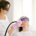 A technician wearing gloves and protective glasses performs a facial laser treatment on a patient lying down, also wearing protective glasses and a hair cap, addressing various skin concerns with advanced laser treatments.
