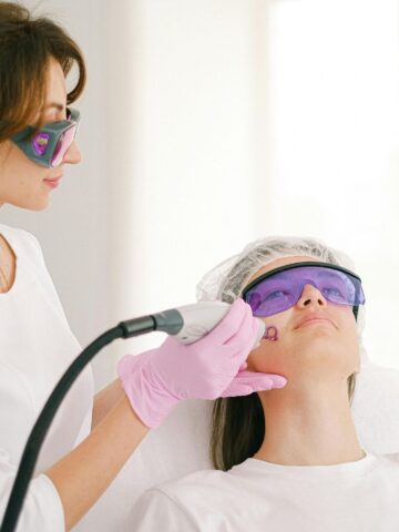 Top 6 Skin Concerns That Laser Treatments Can Address A technician wearing gloves and protective glasses performs a facial laser treatment on a patient lying down, also wearing protective glasses and a hair cap, addressing various skin concerns with advanced laser treatments.