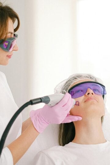A technician wearing gloves and protective glasses performs a facial laser treatment on a patient lying down, also wearing protective glasses and a hair cap, addressing various skin concerns with advanced laser treatments.