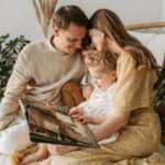 A family of four sits closely together on a bed, smiling and looking at an open book of wedding photos; two children are seated between the adults, cherishing memories as couples often do.