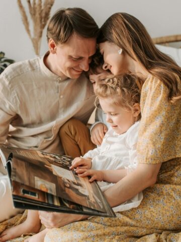A family of four sits closely together on a bed, smiling and looking at an open book of wedding photos; two children are seated between the adults, cherishing memories as couples often do.