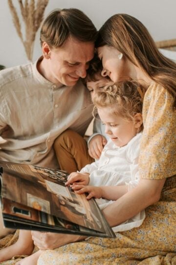 A family of four sits closely together on a bed, smiling and looking at an open book of wedding photos; two children are seated between the adults, cherishing memories as couples often do.