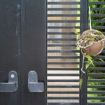 Black aluminium gate with horizontal slats, two silver handles, and a hanging brown pot containing dried plants and some green leaves-combining security with modern style to enhance your property appearance.