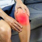 A person sitting on a sofa holds their knee, which appears red and inflamed, suggesting pain or swelling from a possible meniscus tear.