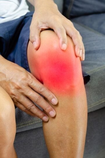 A person sitting on a sofa holds their knee, which appears red and inflamed, suggesting pain or swelling from a possible meniscus tear.