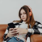 Teenage girl wearing orange headphones sits on a sofa, looking at her mobile with a neutral expression-capturing the quiet moments of a struggling teen potentially seeking private support outside the NHS.
