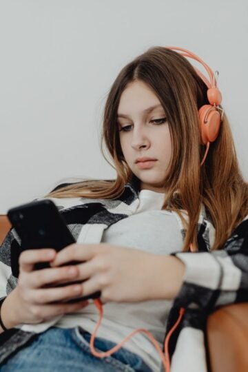 Teenage girl wearing orange headphones sits on a sofa, looking at her mobile with a neutral expression—capturing the quiet moments of a struggling teen potentially seeking private support outside the NHS.