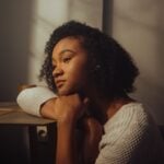 A woman with curly hair rests her chin on her arms whilst sitting at a table, gazing thoughtfully into the distance in soft indoor lighting, reflecting on everyday coping skills for real-life situations.
