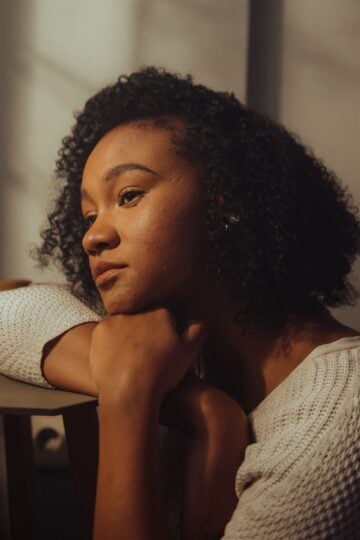 A woman with curly hair rests her chin on her arms whilst sitting at a table, gazing thoughtfully into the distance in soft indoor lighting, reflecting on everyday coping skills for real-life situations.