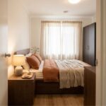 A neatly made bed with brown and beige bedding, a wooden bedside table with a lamp and decor, a wardrobe, and sheer curtains over a window show the cosy results of this budget bedroom makeover in a softly lit room.