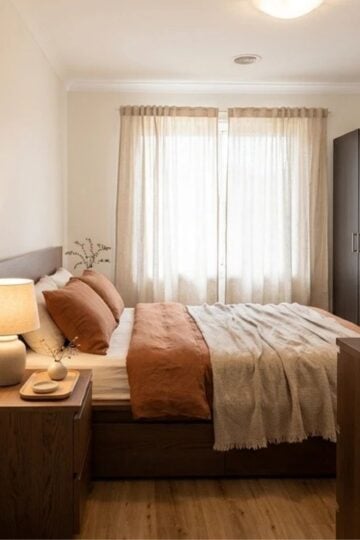 A neatly made bed with brown and beige bedding, a wooden bedside table with a lamp and decor, a wardrobe, and sheer curtains over a window show the cosy results of this budget bedroom makeover in a softly lit room.