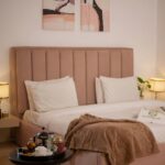 A neatly made bed with white linen, a light brown headboard, beige blanket, and bedside tables with lamps creates a calm bedroom vibe; a tray with tea set completes this inviting family bedroom interior design.