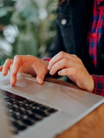 A person types on a laptop at a wooden table, perhaps undertaking a cybersecurity course. They wear a red checked shirt under a dark jacket. The background is blurred with green plants visible.