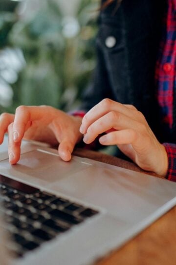 A person types on a laptop at a wooden table, perhaps undertaking a cybersecurity course. They wear a red checked shirt under a dark jacket. The background is blurred with green plants visible.