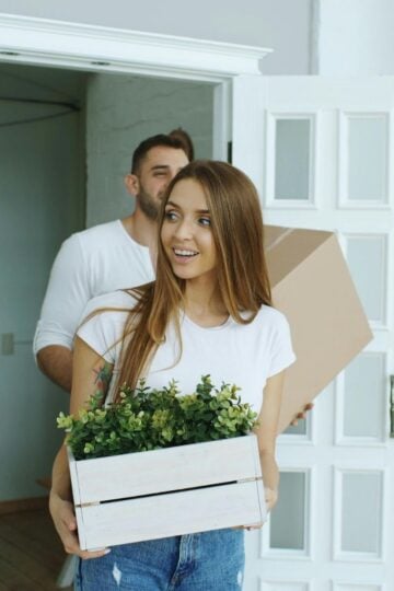 Two people enter a room; the woman in front carries a white wooden box with green plants, whilst the man behind, feeling the usual moving challenges, carries a cardboard box on this hectic moving day.