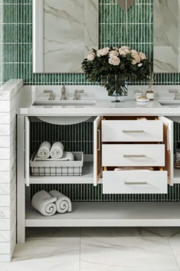 Modern family bathroom with a white, durable vanity featuring double basins, open drawers and shelves displaying towels and baskets, a green tiled wall, and a vase of flowers on the worktop.