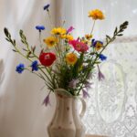 A ceramic vase with a floral design holds a bouquet of colourful wildflowers, including yellow, red, blue, and purple blooms-each arranged to deliver freshness-placed on a table near a lace-curtained window.