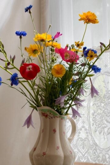 A ceramic vase with a floral design holds a bouquet of colourful wildflowers, including yellow, red, blue, and purple blooms—each arranged to deliver freshness—placed on a table near a lace-curtained window.