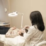 A person in a lab coat and gloves performs a facial procedure on a patient lying on a treatment couch under a bright examination lamp.