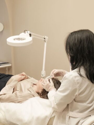 A person in a lab coat and gloves performs a facial procedure on a patient lying on a treatment couch under a bright examination lamp.
