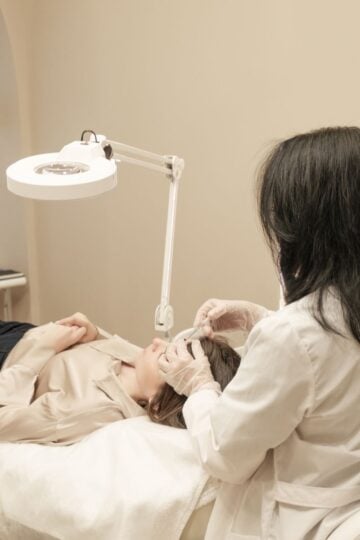 A person in a lab coat and gloves performs a facial procedure on a patient lying on a treatment couch under a bright examination lamp.