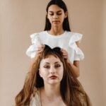 A young woman stands behind another woman, gently combing her long brown hair, as if preparing to style it with one of the latest lightweight wigs, against a plain beige background.