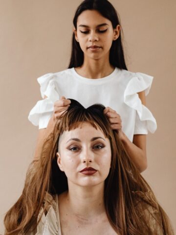 A young woman stands behind another woman, gently combing her long brown hair, as if preparing to style it with one of the latest lightweight wigs, against a plain beige background.