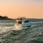 A white motor yacht, perfect for luxury travel or a private yacht charter, moves through the water at sunset, leaving a wake behind. Trees line the left shore.