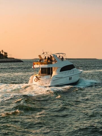 A white motor yacht, perfect for luxury travel or a private yacht charter, moves through the water at sunset, leaving a wake behind. Trees line the left shore.