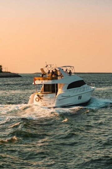 A white motor yacht, perfect for luxury travel or a private yacht charter, moves through the water at sunset, leaving a wake behind. Trees line the left shore.
