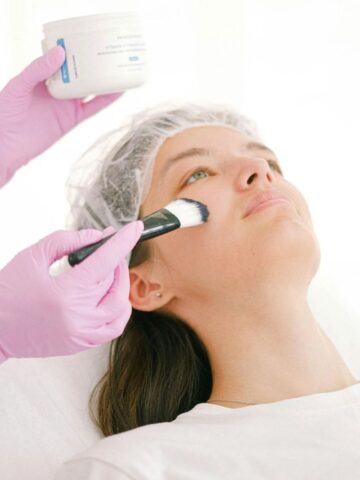 A person wearing a hair cap receives non-surgical facial enhancements as another person in pink gloves applies a cream to their face with a brush.