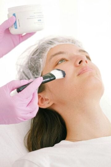 A person wearing a hair cap receives non-surgical facial enhancements as another person in pink gloves applies a cream to their face with a brush.