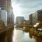 Urban canal flanked by modern high-rise buildings on both sides, with a footbridge crossing over the water and sunlight shining from the left-a perfect scene showcasing reasons to move to Manchester.