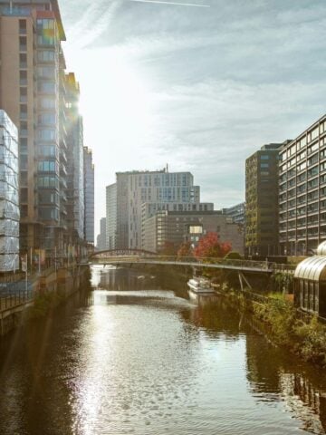 Urban canal flanked by modern high-rise buildings on both sides, with a footbridge crossing over the water and sunlight shining from the left-a perfect scene showcasing reasons to move to Manchester.