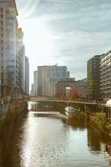 Urban canal flanked by modern high-rise buildings on both sides, with a footbridge crossing over the water and sunlight shining from the left—a perfect scene showcasing reasons to move to Manchester.