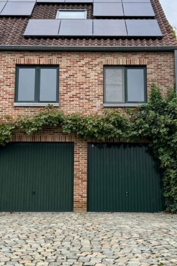 Two green garage doors on a brick house with solar panels on the roof and vines growing around the doors. Built by expert builders to meet Future Homes Standard building regulations. Cobbled driveway in front.