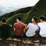 Four young men sit on the edge of a hill, facing away, overlooking green mountains and a distant valley under a cloudy sky-an inspiring moment reflecting the importance of men's health and connection with nature.