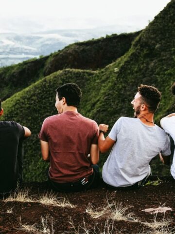Four young men sit on the edge of a hill, facing away, overlooking green mountains and a distant valley under a cloudy sky-an inspiring moment reflecting the importance of men's health and connection with nature.