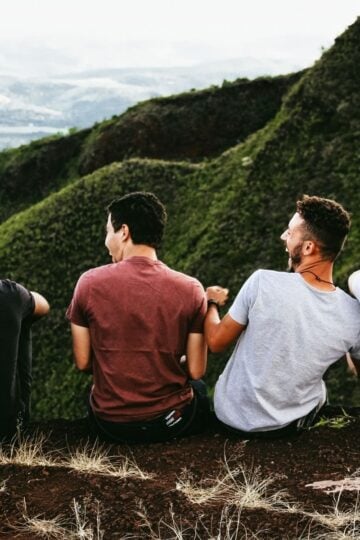 Four young men sit on the edge of a hill, facing away, overlooking green mountains and a distant valley under a cloudy sky—an inspiring moment reflecting the importance of men’s health and connection with nature.