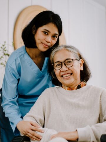 A young woman in blue scrubs stands next to an older woman in a wheelchair, both smiling at the camera inside a well-lit room, reflecting carer companionship and warmth.