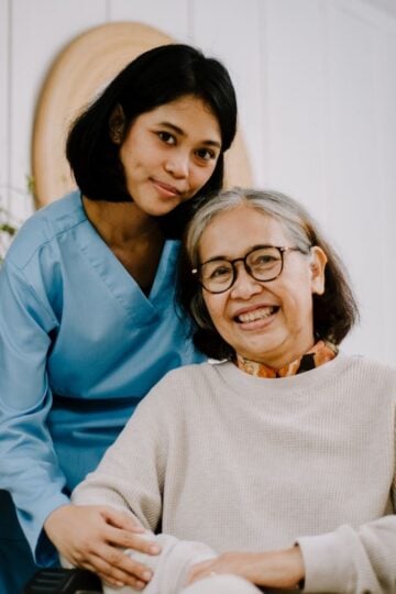 A young woman in blue scrubs stands next to an older woman in a wheelchair, both smiling at the camera inside a well-lit room, reflecting carer companionship and warmth.