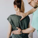 A healthcare professional examines a woman's upper back and neck, checking for spine symptoms while she sits facing away from the camera.